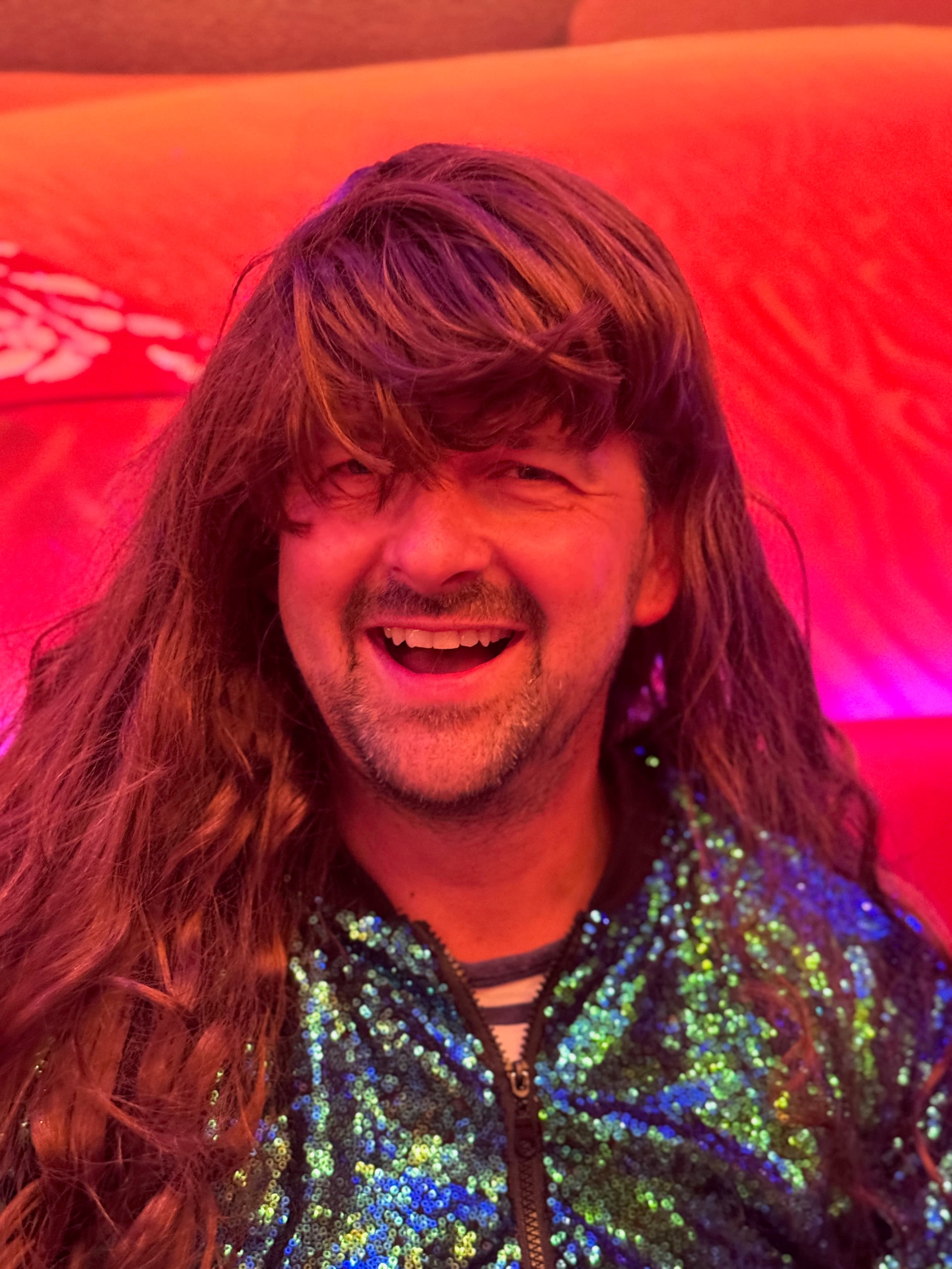 Smiling guest in a long brunette wig and sequined jacket under red lighting